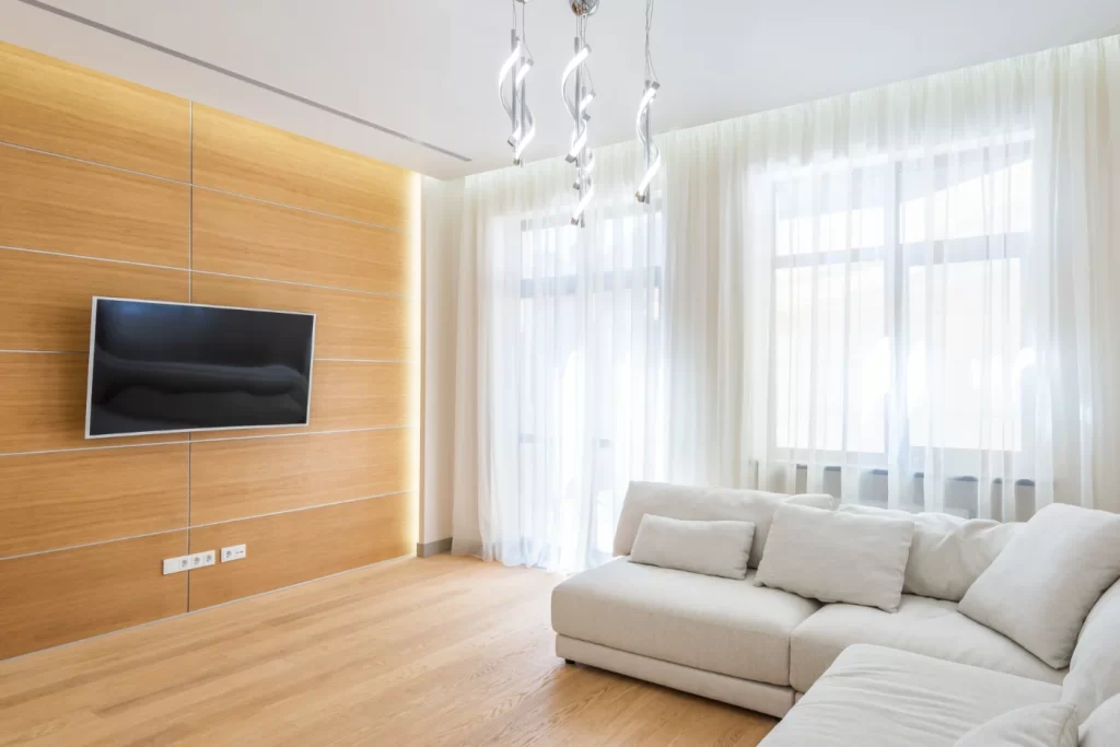 Home remodeling Modern living room featuring a minimalist design with a sleek wall-mounted TV, light wood paneling, a cozy white sectional sofa, and large windows letting in natural light.
