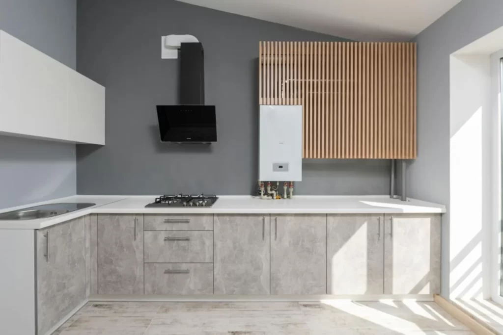 Modern kitchen remodeling with minimalist design, featuring light gray cabinets, white countertops, wall-mounted black range hood, and wooden slatted panel above a white boiler on a gray wall background.